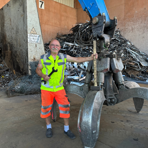 Mitarbeiter mit Greifarm in Schrotthalle Arbeiter in Warnkleidung steht vor einem großen Greifarm einer Baumaschine in einer Halle mit Metallschrott im Hintergrund.